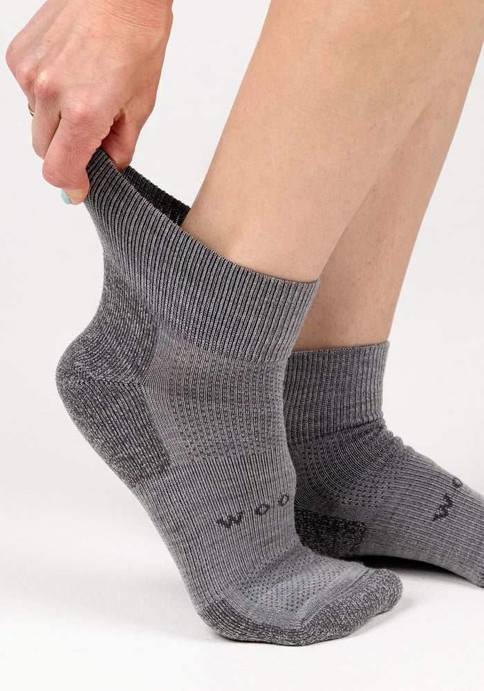 A person pulls on a Woolx Womens Rambler Ankle Sock - Pebble with their right hand, already wearing the matching gray sock. These Merino wool socks have a subtle ribbed pattern and provide great moisture management against a white background.