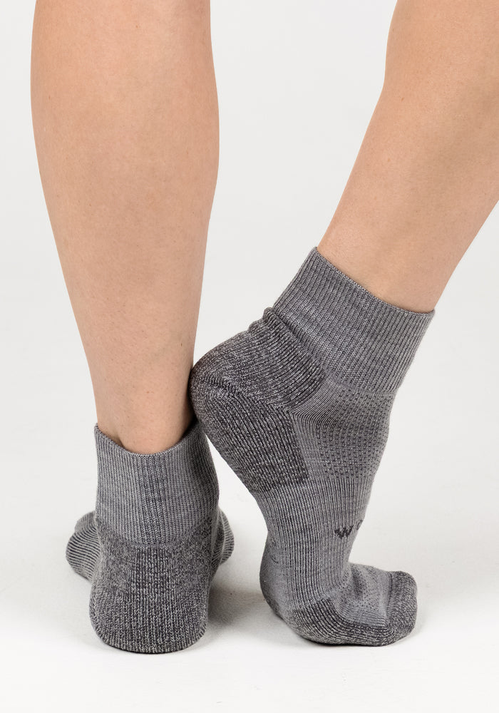 Two feet wearing Woolx Womens Rambler Ankle Sock - Pebble stand on a white background, one flat and one crossed behind, showing back and side views while highlighting excellent moisture management.