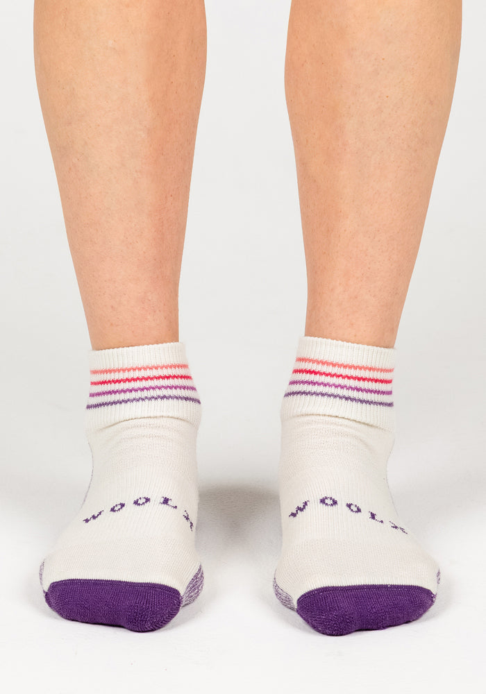 A pair of feet wear Woolx Womens Rambler Ankle Socks in Sand Dollar—white Merino wool with purple toes, heels, and red and purple striped cuffs. "WOOL" in purple highlights these moisture-wicking socks.
