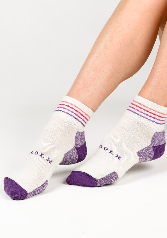 A person wears Woolx Womens Rambler Ankle Sock - Sand Dollar, featuring moisture-wicking white fabric, purple toes and heels, red and pink cuff stripes, and "COOL" on top. Their legs rest comfortably on a white surface.