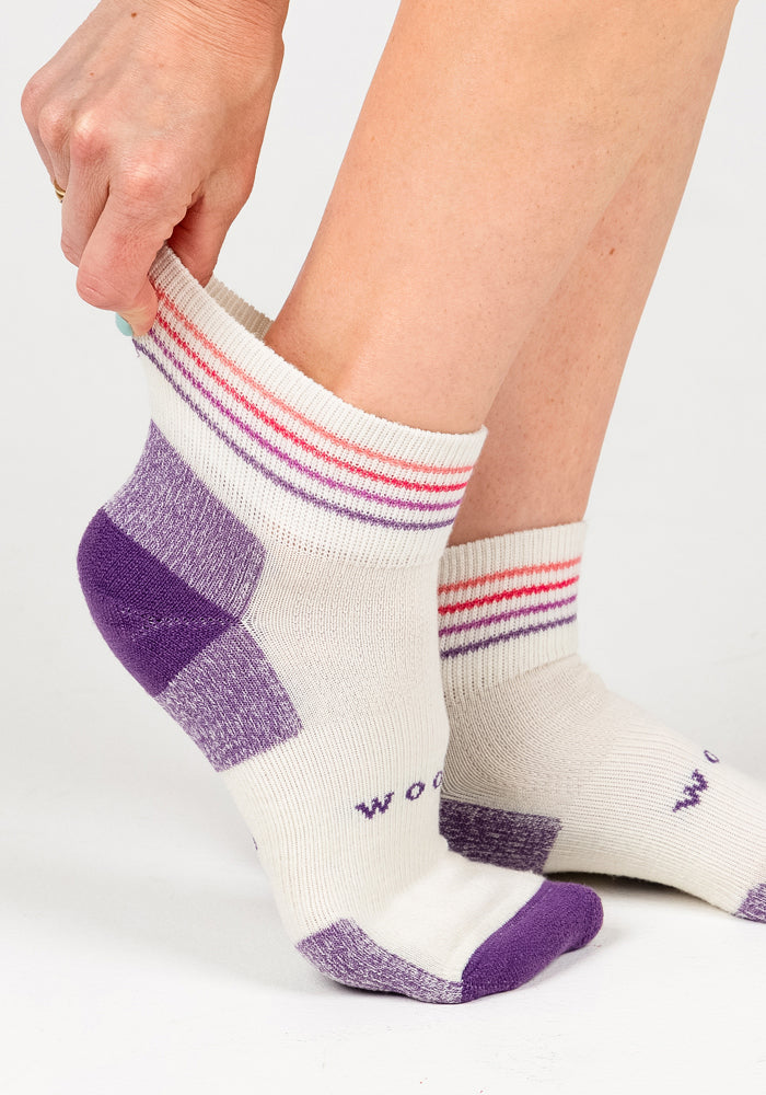 A person pulls on a Woolx Womens Rambler Ankle Sock - Sand Dollar, featuring white Merino wool with purple toes, heels, and pink stripes. The soft, moisture-wicking socks are shown on lower legs and feet.