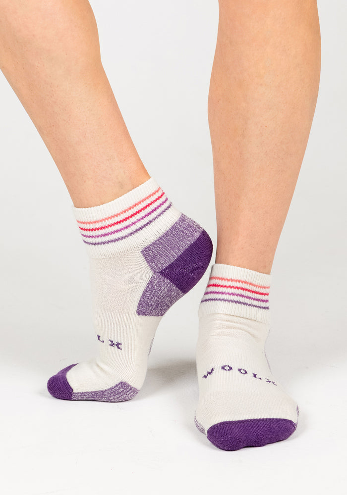 A person wears Woolx Womens Rambler Ankle Socks in Sand Dollar, featuring purple toes, heels, and soles with red-striped cuffs. "WOOLX" is on the bottoms. The moisture-wicking socks are shown against a plain white background.