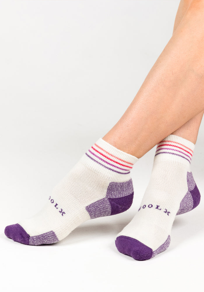 A person wears the Woolx Womens Rambler Ankle Sock - Sand Dollar, featuring white Merino wool with purple toes, heels, and accents, plus pink and purple cuff stripes. "WOOLX" is printed in purple on the side for stylish comfort.