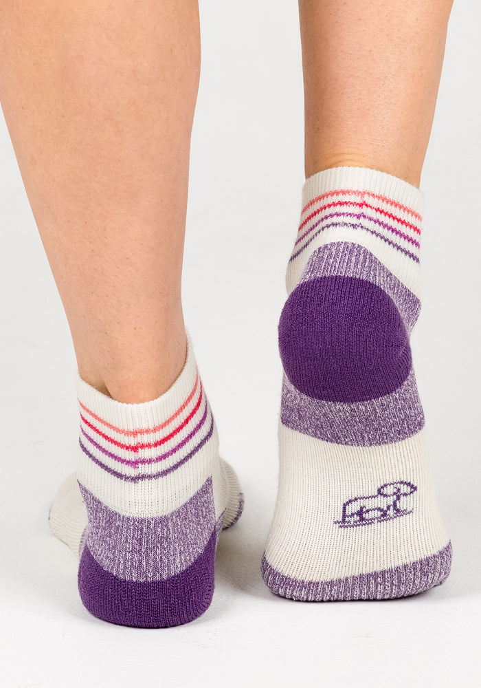 A close-up of two feet wearing Woolx Womens Rambler Ankle Sock - Sand Dollar in white with purple accents and logo, set against a plain white background.