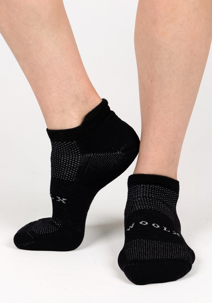 A close-up of two feet wearing Woolx Womens Rowan No Show Socks in Black Pepper, featuring white dotted patterns and text, posed against a plain white background.