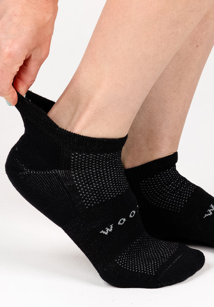 A person pulls on a Woolx Womens Rowan No Show Sock in Black Pepper, featuring cushioning, a textured pattern, and moisture-wicking fabric. The sock is worn on a bare foot against a plain white background.