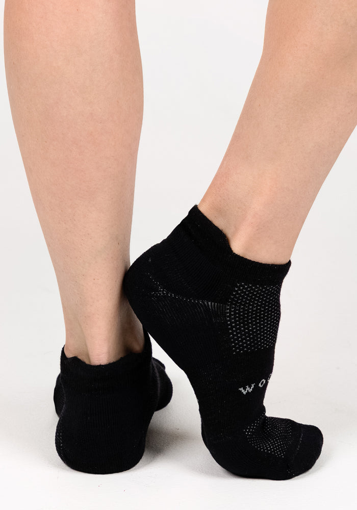 Close-up of a person wearing Woolx Womens Rowan No Show Socks in Black Pepper, standing on a white surface with one foot resting on its toes behind the other leg.