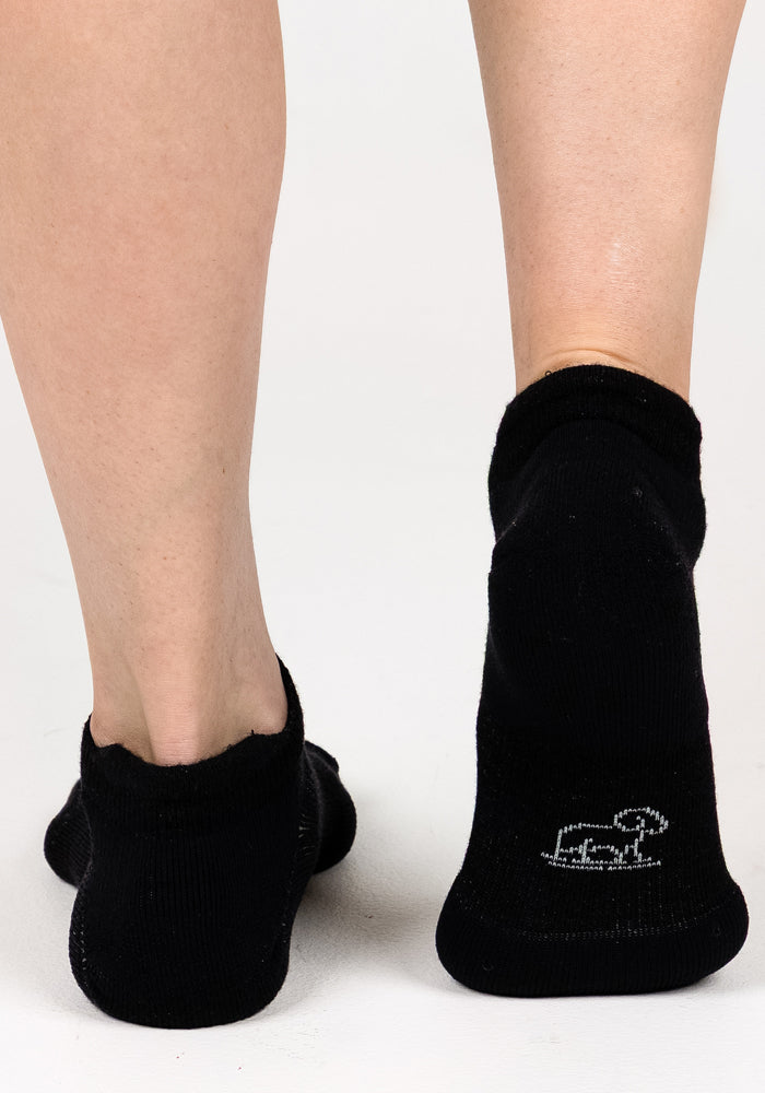 A close-up of two feet in Woolx Womens Rowan No Show Socks - Black Pepper, standing on a white surface; one sock has a small white embroidered design near the heel.