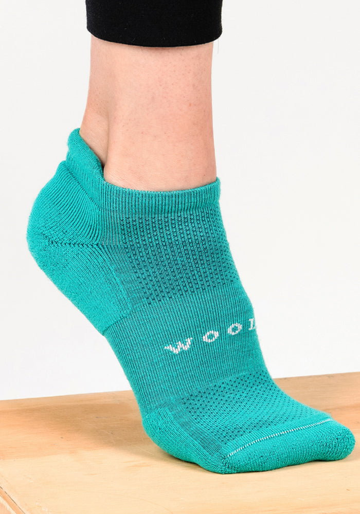 A person wearing Woolx Womens Rowan No Show Socks in Cool Jade stands on a wooden surface. The breathable Merino wool sock has ribbed details and "WOOL" partially visible in white letters on the side.