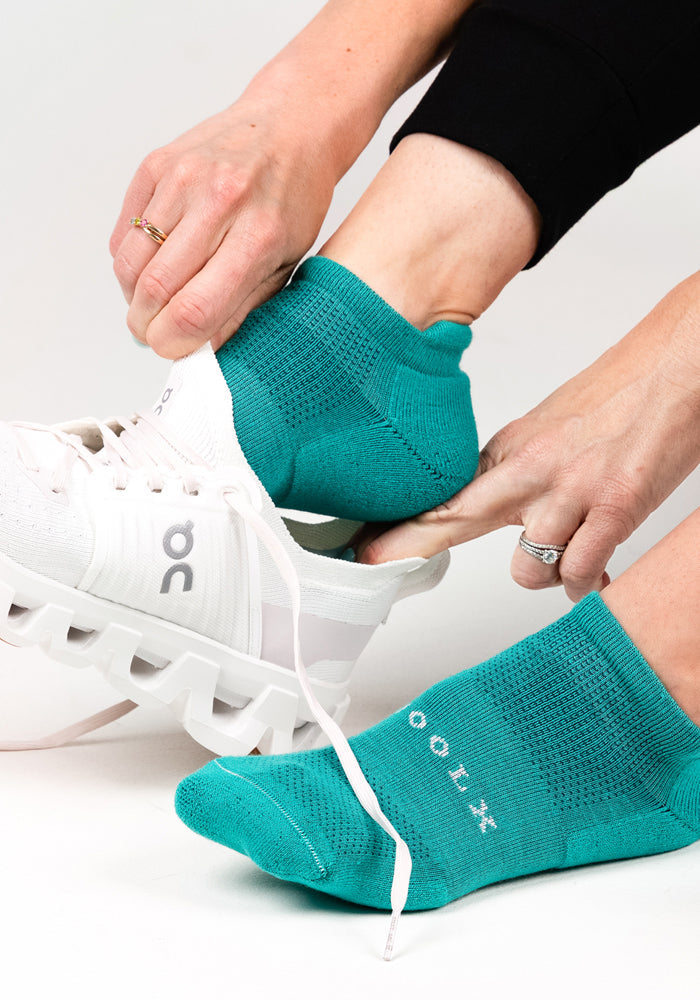 Wearing Woolx Womens Rowan No Show Socks in Cool Jade and white athletic shoes, a person adjusts their shoe while the other foot in the same socks rests on the ground. Both hands with rings are visible.