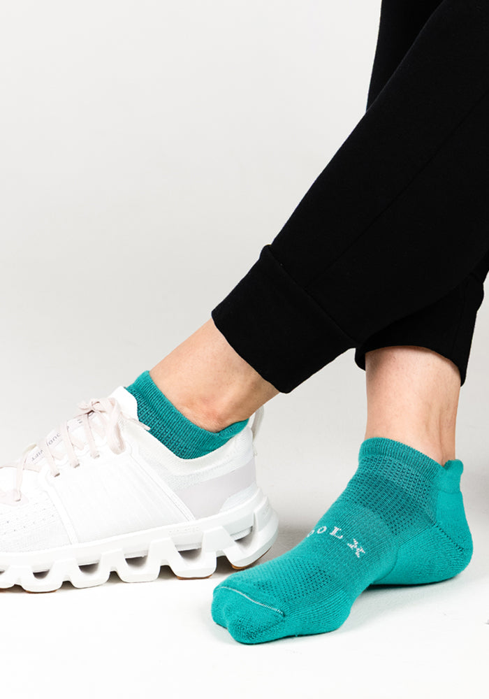 A person wears black pants and Woolx Womens Rowan No Show Socks in Cool Jade, with one white sneaker on and the other foot displaying the sock’s white text. The plain white background highlights the moisture-wicking Merino wool socks.