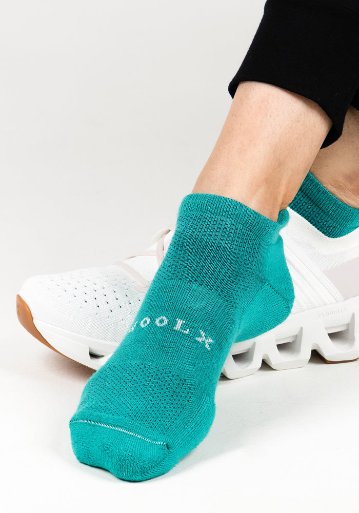 A woman wears Woolx Womens Rowan No Show Socks in Cool Jade, featuring white text, paired with white athletic shoes and sitting cross-legged on a white background.