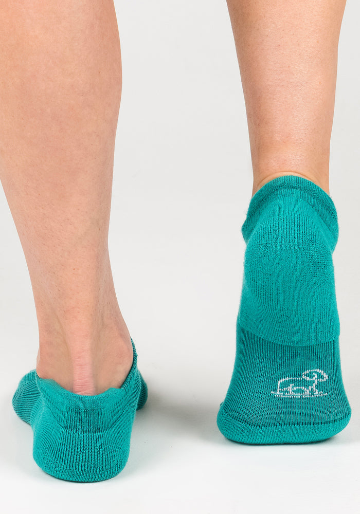 A person in Woolx Womens Rowan No Show Socks - Cool Jade stands on a white surface, one foot ahead. The right sock displays a small white logo on the heel, emphasizing their moisture-wicking comfort.