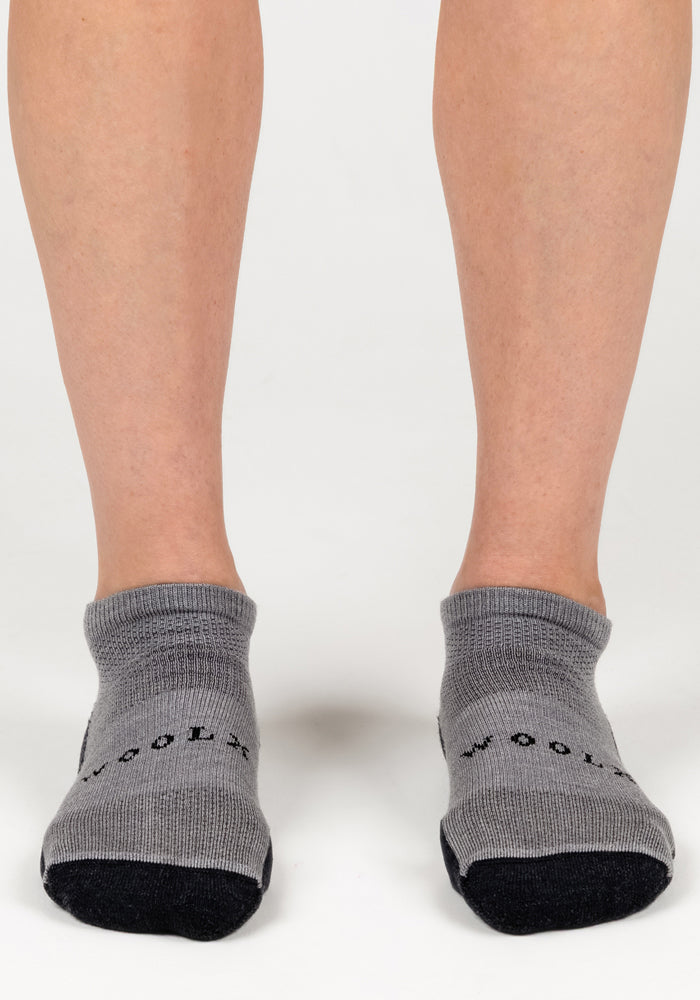 A pair of feet wears Woolx Womens Rowan No Show Socks in Pebble on a white background. "WOOL" appears near the toes, highlighting these cozy Merino wool socks designed for ultimate comfort.