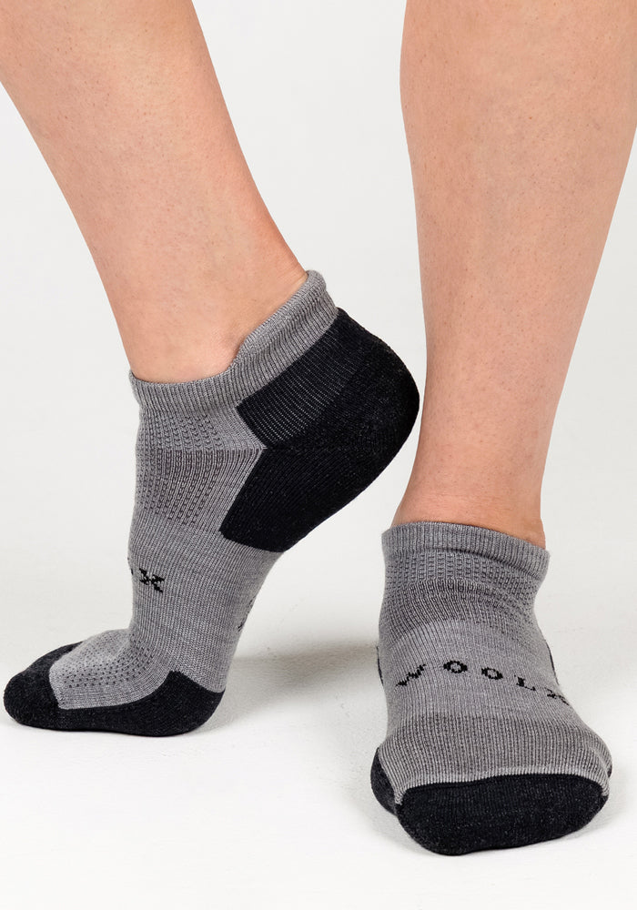 A close-up of two feet wearing Woolx Womens Rowan No Show Socks in Pebble, featuring black heels and toes, small black text, breathable fabric, and reinforced areas for comfort and durability on a white surface.