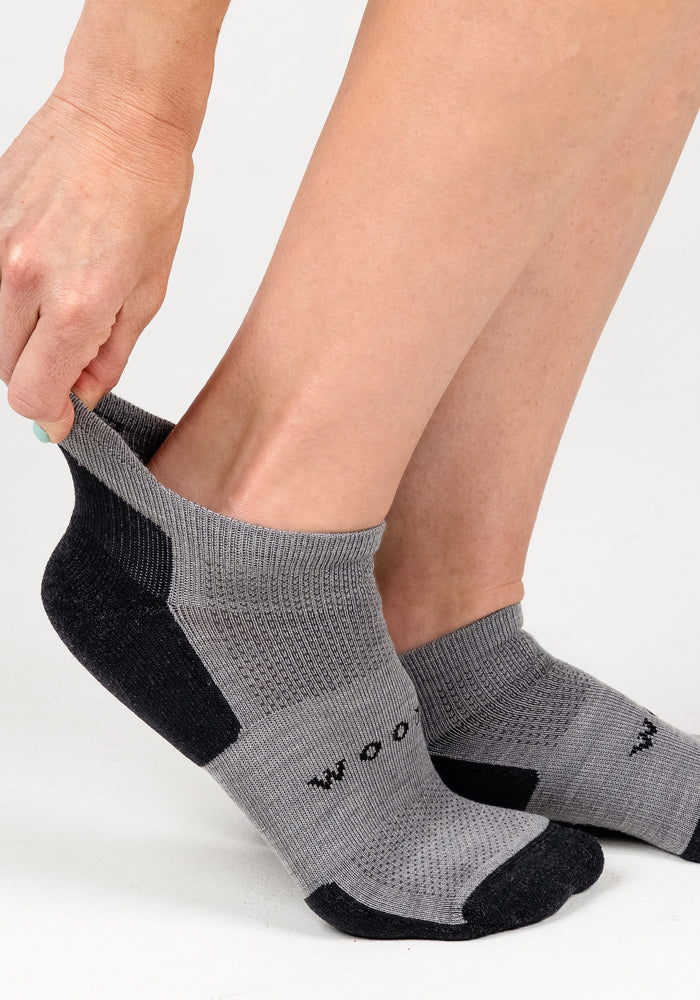 A person pulls on Woolx Womens Rowan No Show Socks in Pebble, featuring gray and black Merino wool, breathable textured fabric, and "WOOL" on the side, against a plain white background.