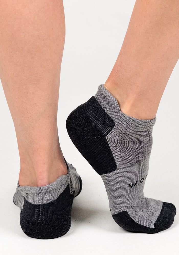 Close-up of a person wearing Woolx Womens Rowan No Show Socks in Pebble, standing on a light surface with one foot flat and the other raised, highlighting the comfort and breathability of these gray and black socks.