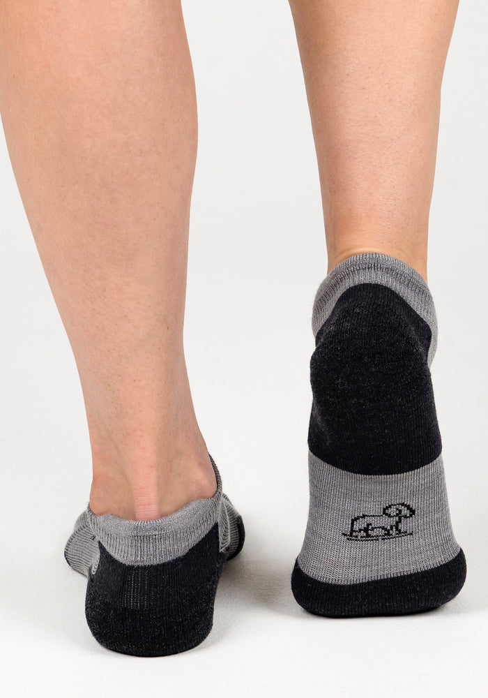 A close-up shows a person’s lower legs and feet in Woolx Womens Rowan No Show Socks - Pebble, featuring cushioned comfort, a black and gray design, and a small dog graphic on the back against a plain white background.