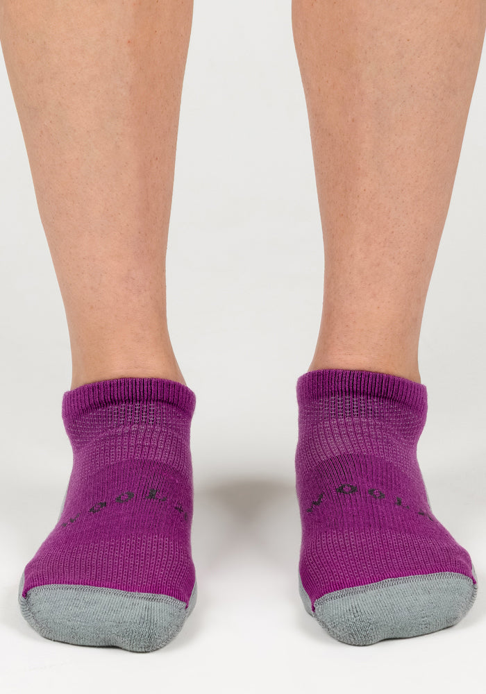 A pair of feet wears Woolx Womens Rowan No Show Socks in Purple Wine with gray toes, standing on a white surface.