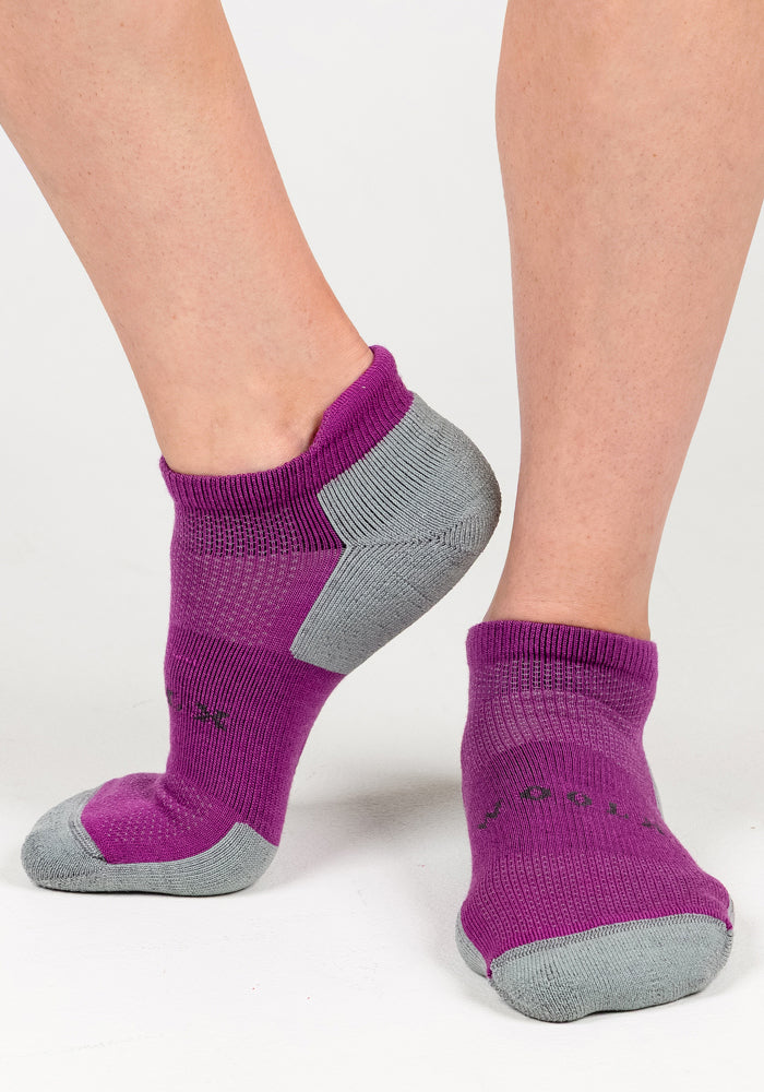 A close-up shows two feet wearing Woolx Womens Rowan No Show Socks in Purple Wine, featuring gray heels, toes, and soles, posed on a white background.