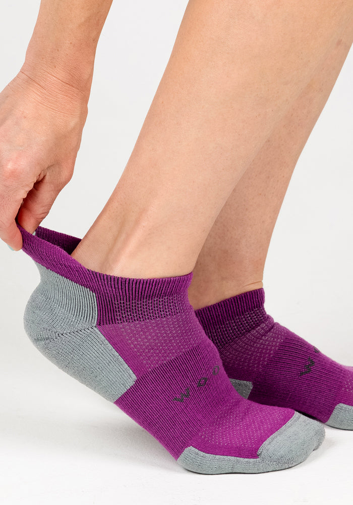 A woman puts on a Woolx Womens Rowan No Show Sock in Purple Wine, using one hand to hold the heel with her foot partially inside. The background is plain white.