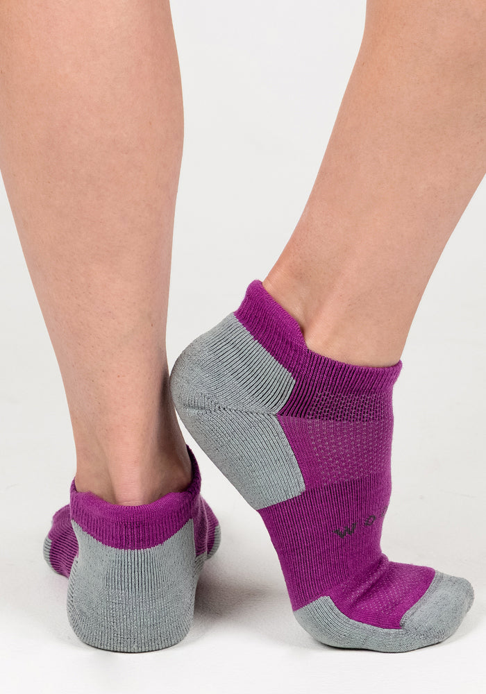A pair of feet wear Woolx Womens Rowan No Show Socks in Purple Wine, featuring cushioned ankle support with gray heels and toes. One foot is flat and the other slightly raised, highlighting both back and side views against a white background.