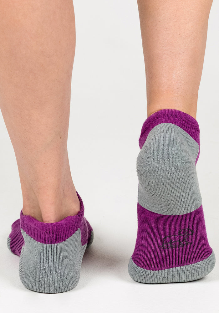 Close-up of a person’s lower legs wearing Woolx Womens Rowan No Show Socks in Purple Wine. The cushioned, breathable socks feature a small logo on the heel and are shown from the back on a white background.