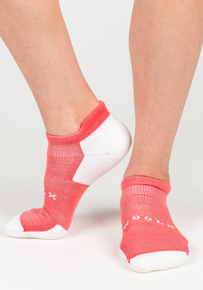A person wears Woolx Womens Rowan No Show Socks - Taffy Twist in pink and white, standing on tiptoe with one foot raised to showcase the fit and design against a neutral background.