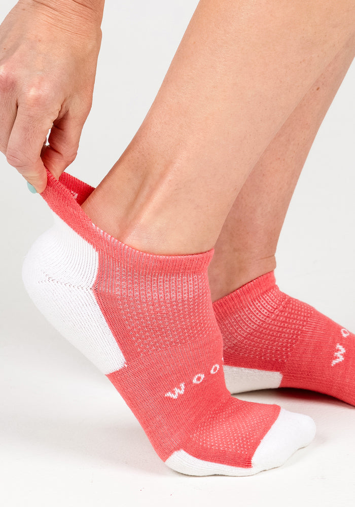 A person adjusts a Womens Rowan No Show Sock - Taffy Twist by Woolx on their foot, with a plain, light background highlighting the pink and white cushioned sock and hand.