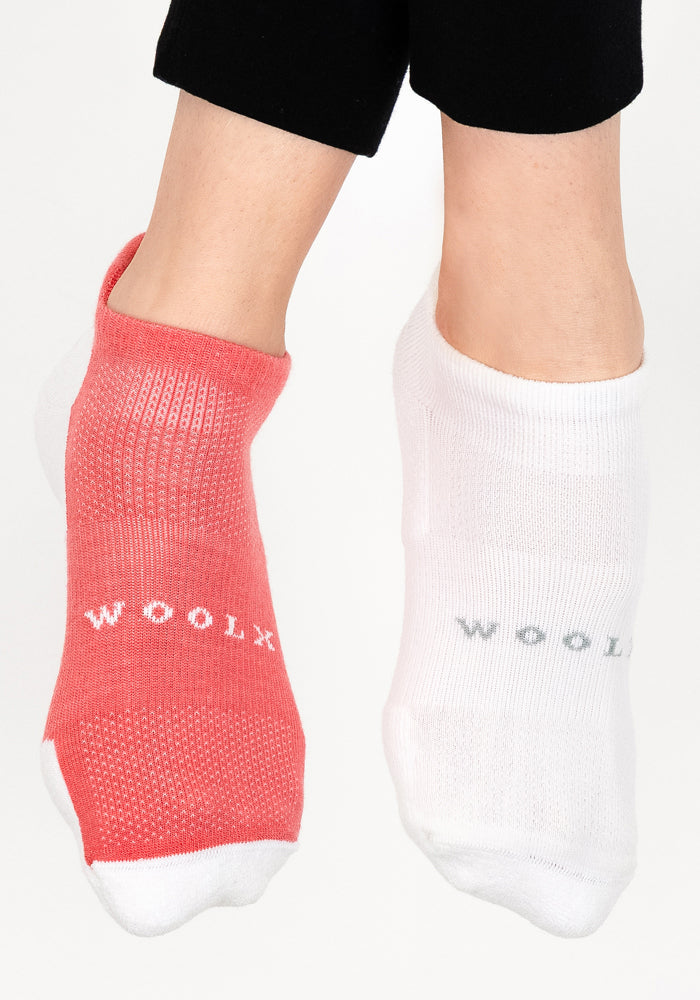 A person wears mismatched Woolx Womens Rowan No Show Socks in Taffy Twist—one coral red, one white—with black pants, highlighting the cushioned Merino wool comfort against a white background.