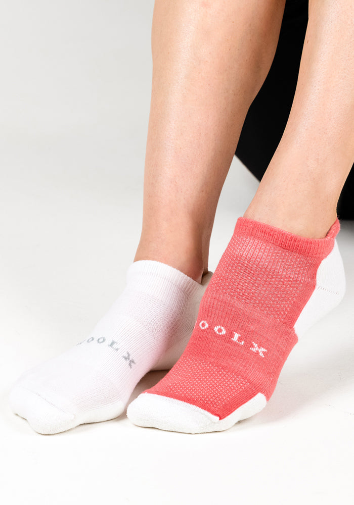 A person wears Woolx Womens Rowan No Show Socks in Taffy Twist—one white with gray text, one pink with white text—while sitting on a white background with legs slightly crossed.
