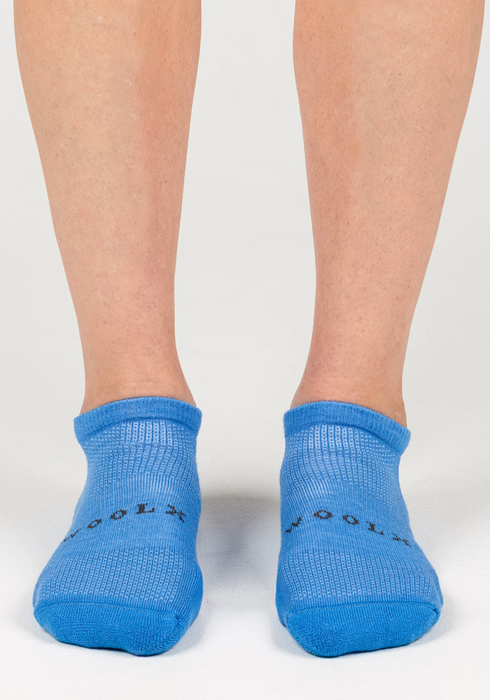 A person wearing Woolx Womens Rowan No Show Socks in Wave Rider, featuring light blue moisture-wicking fabric and black toe text, stands on a light surface. Only their lower legs and feet are visible.