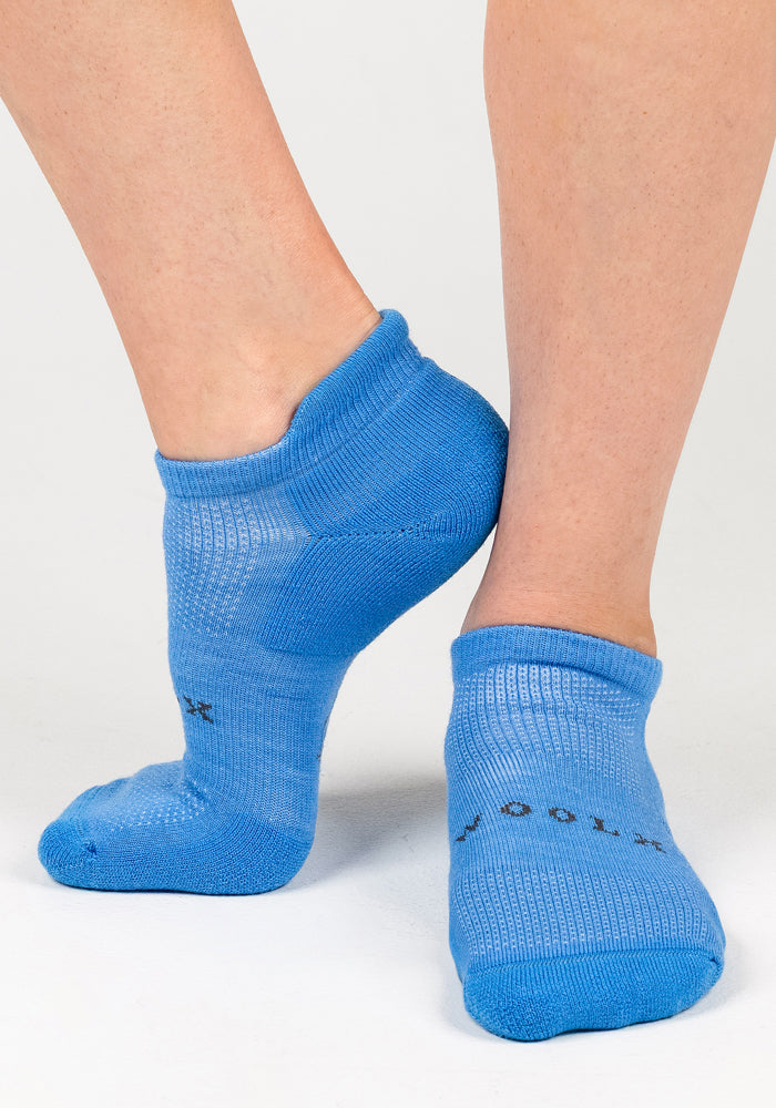 A close-up of two feet in bright blue Womens Rowan No Show Socks - Wave Rider by Woolx. The Merino wool socks have a slight sheen and "WOOLX" in black near the toes, set against a plain white background.