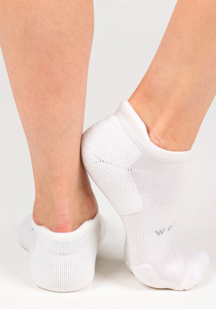 Close-up of two feet wearing Woolx Womens Rowan No Show Socks in White Lightning, one foot on tiptoe and the other flat, shown against a plain white background.
