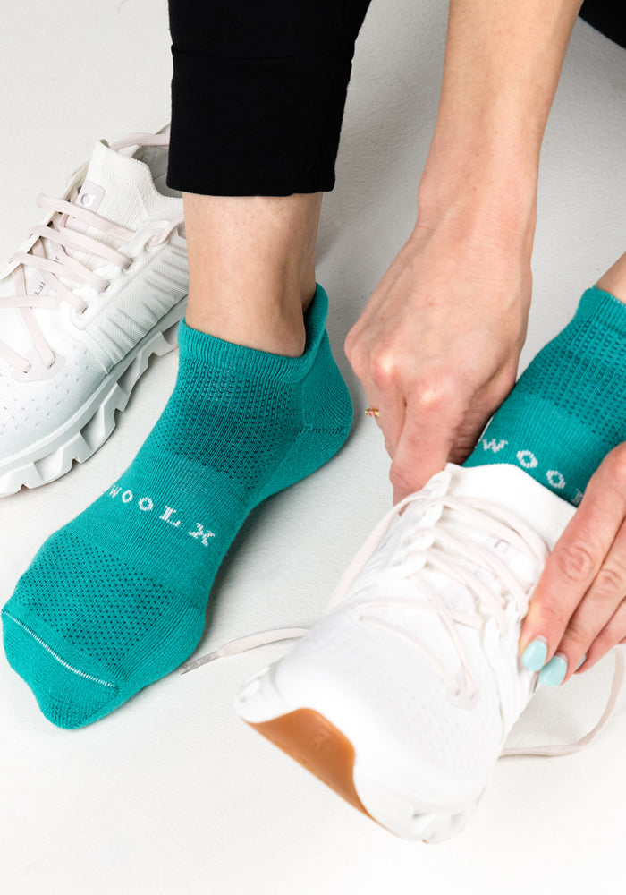 A person wearing Woolx Womens Rowan No Show Socks in Cool Jade and black leggings is putting on a white athletic shoe, while the other foot is already in a shoe, tying the laces.