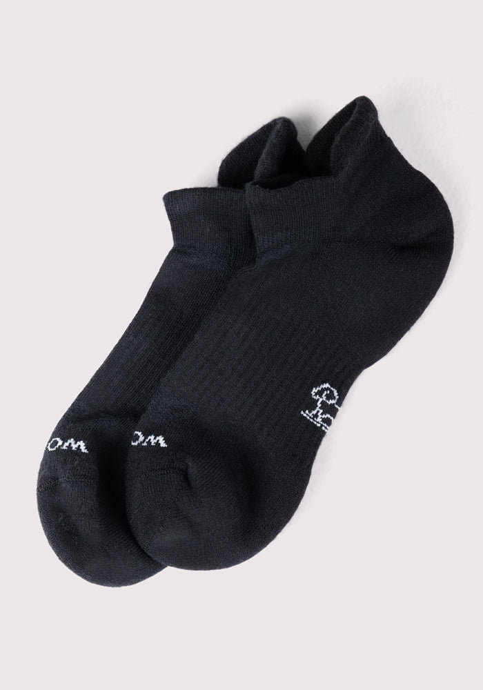 The Woolx Womens Rowan No Show Socks in black feature cushioning, a small white logo and white text near the toes, shown against a light gray background.