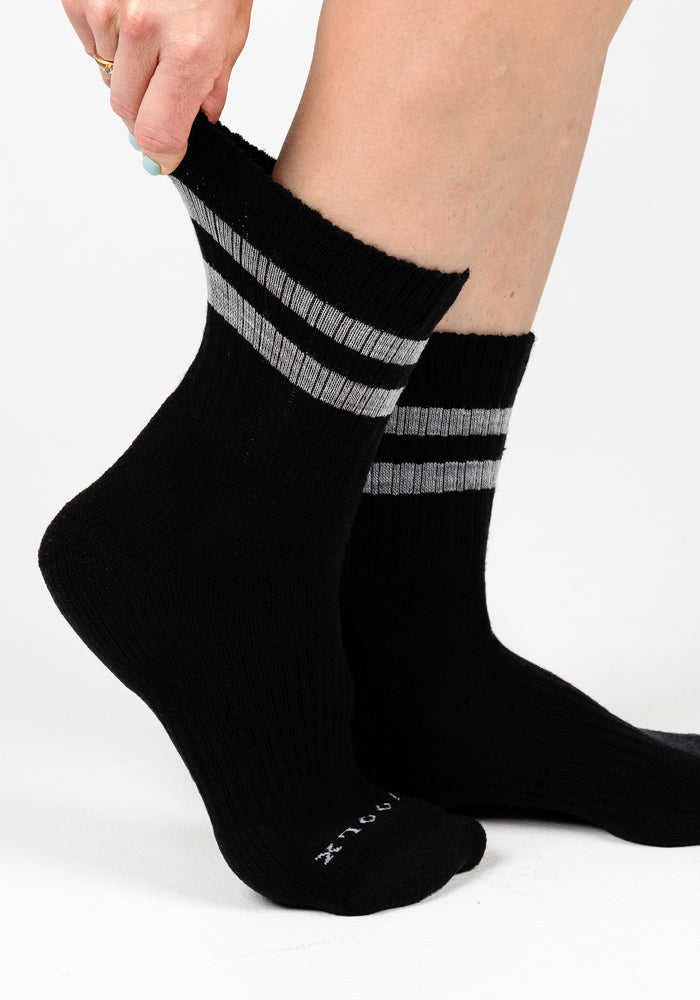 A person wears Woolx Womens Varsity Mini Crew Socks - Black Volt, featuring two gray stripes near the top. Both matching socks are shown against a plain white background.