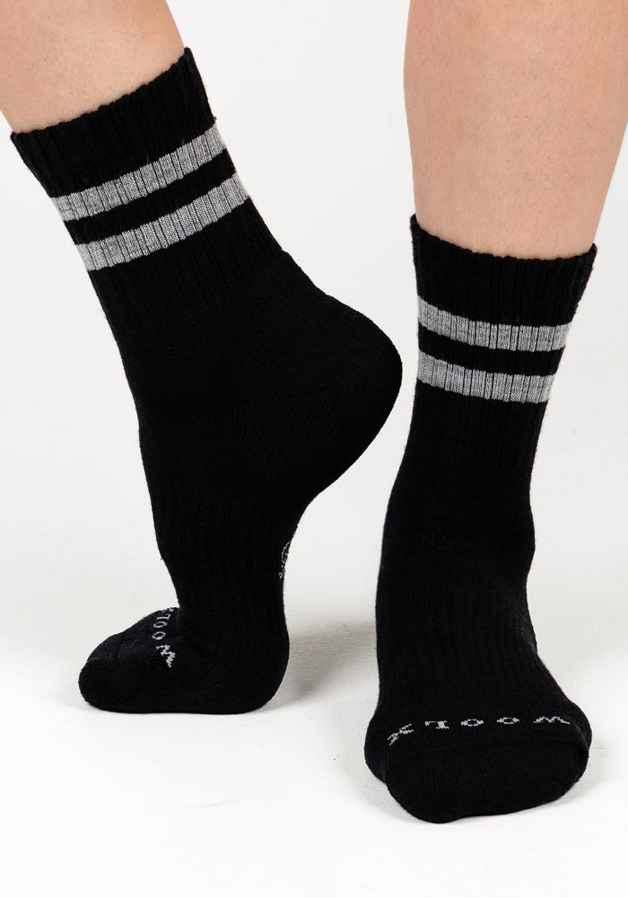 A pair of feet wearing Woolx Womens Varsity Mini Crew Socks - Black Volt, featuring two gray stripes near the top, shown against a plain white background.