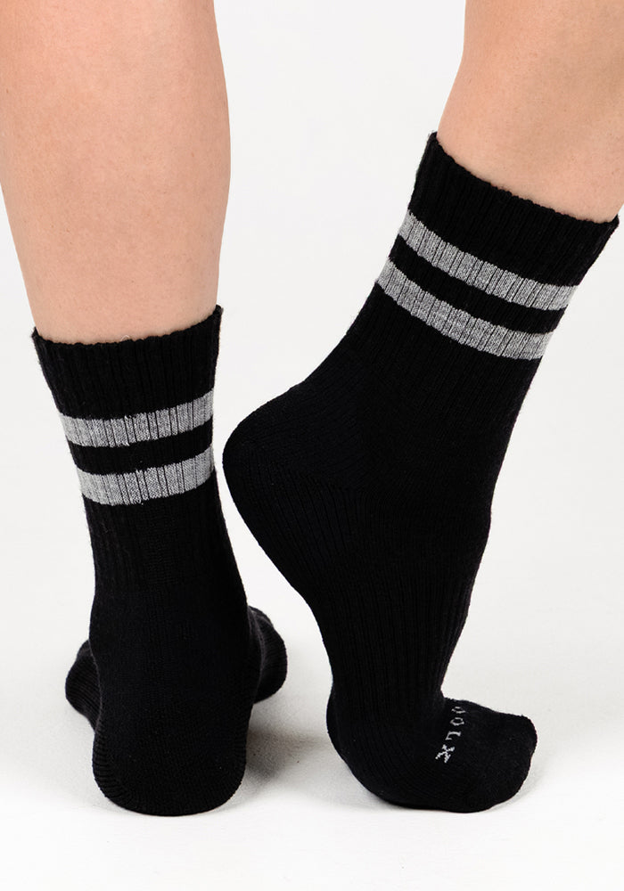 A woman stands on a white surface, heel lifted to reveal Woolx Womens Varsity Mini Crew Socks in Black Volt with two gray stripes near the top, showcasing their moisture-wicking design from the back and side.