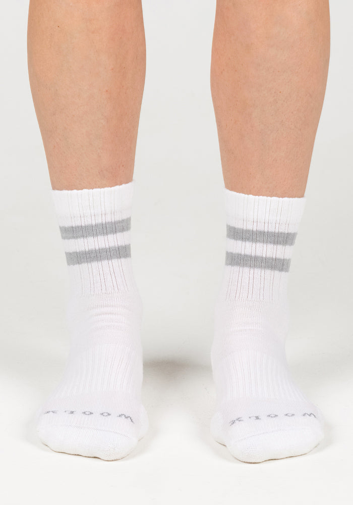 A person stands on a white background wearing Woolx Womens Varsity Mini Crew Socks in Polar White, featuring two gray stripes, ribbed moisture-wicking fabric, and stylish toe text for added comfort.