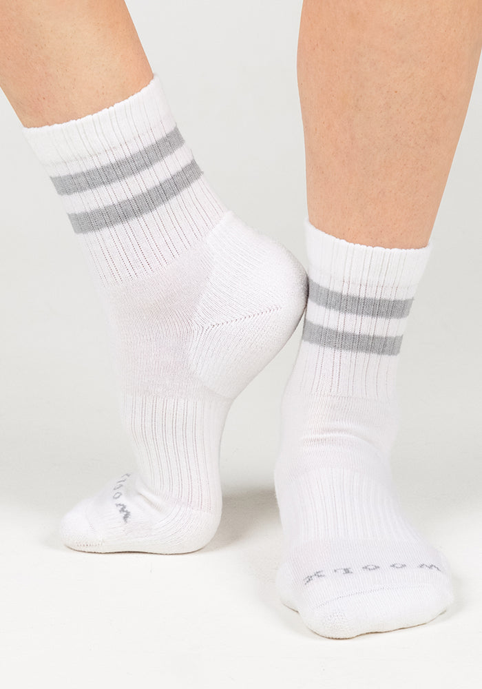 A person stands on tiptoe, showing the side and sole of Woolx Womens Varsity Mini Crew Socks in Polar White with two light gray stripes, highlighting their moisture-wicking design against a plain white background.