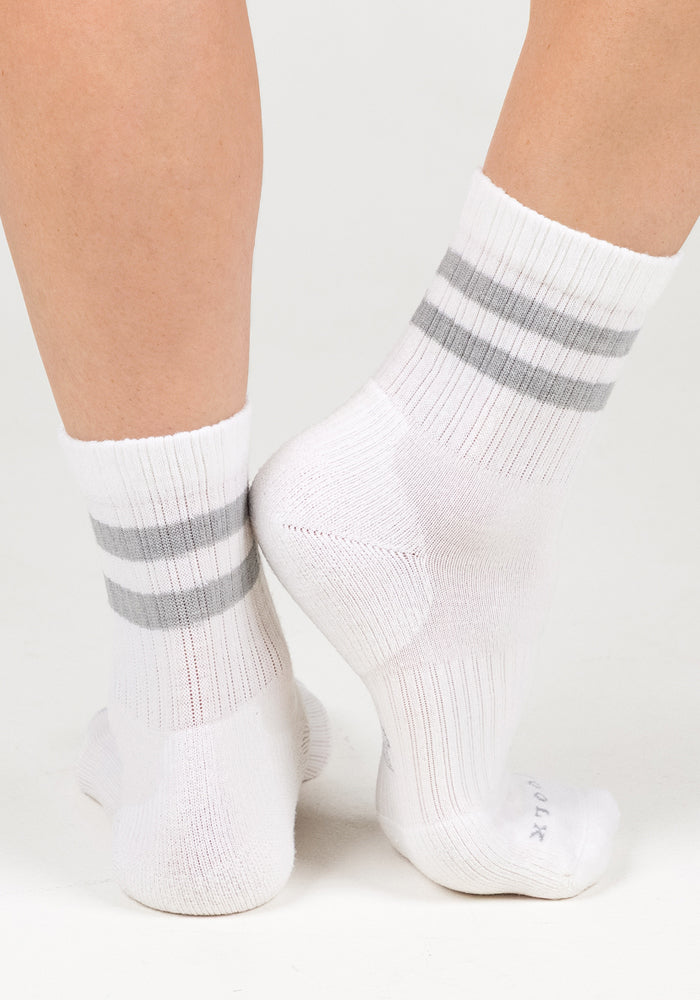 Close-up of two feet wearing Woolx Womens Varsity Mini Crew Socks in Polar White, featuring gray stripes near the top, standing on a white background with one foot crossed behind the other.