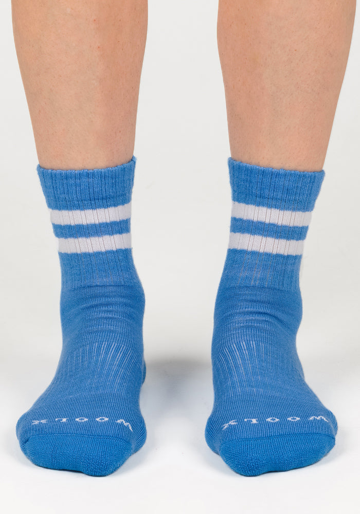 A pair of feet wearing Woolx Womens Varsity Mini Crew Socks - Wave Rider, blue athletic crew socks with two white stripes near the top and small white text on the moisture-wicking toes, standing on a white surface.