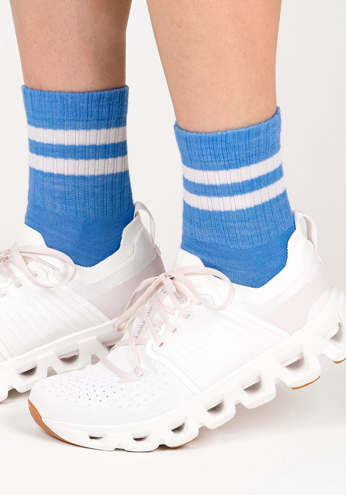 A close-up of two feet in white sneakers and Woolx Womens Varsity Mini Crew Socks - Wave Rider, blue crew socks with white stripes, standing on a white surface.