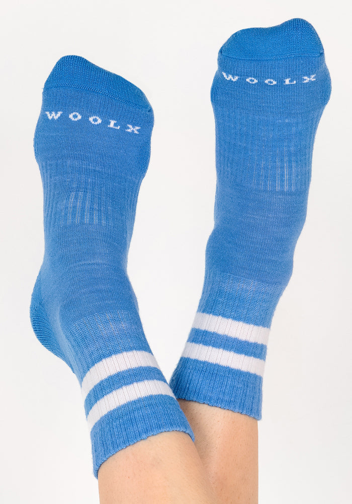 A pair of feet wearing Woolx Womens Varsity Mini Crew Socks - Wave Rider, featuring blue fabric with white stripes near the top and "WOOLX" printed in white on the toes, set against a light background.