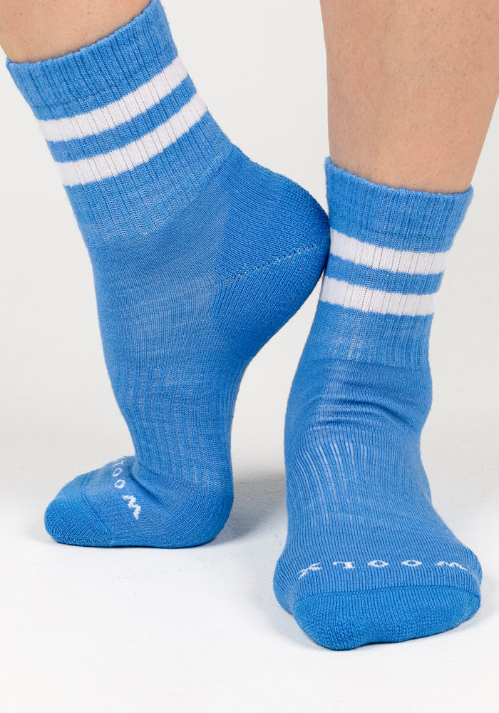 Close-up of two feet in Woolx Womens Varsity Mini Crew Socks - Wave Rider, bright blue with white stripes, standing on a white background. One foot points down while the other is flat, displaying these moisture-wicking socks in detail.
