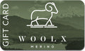 The Woolx Gift Card showcases a minimalist design of a ram on a hill with "WOOLX" and "MERINO" beneath, all set against a scenic mountain backdrop. The card is primarily green and adorned with clouds.