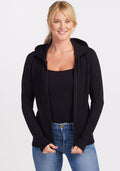A person with long blonde hair is smiling while wearing the Woolx Black Zoey Zip Up Hoodie over a black top made of Australian Merino Wool and light blue jeans. The individual is posing with hands resting on the hips against a plain white background, showcasing perfect layering. | Karly is 5'10", wearing a size S