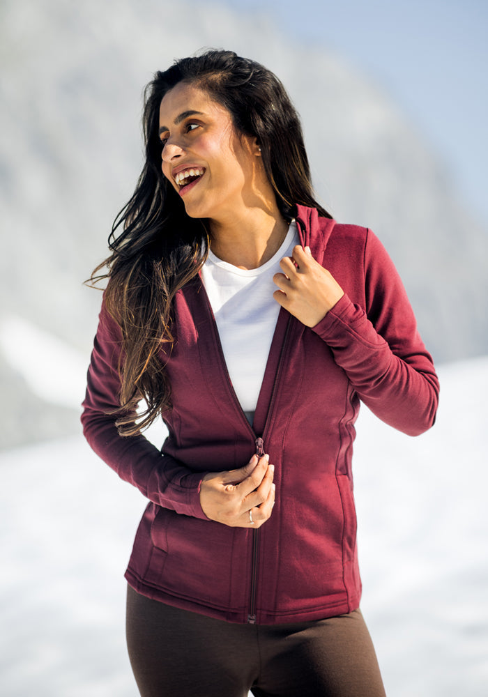 A woman wears a black Woolx Zoey Zip Up Hoodie and brown pants while outdoors in snowy mountains, smiling and looking to the side with her hand on the hoodie zipper.