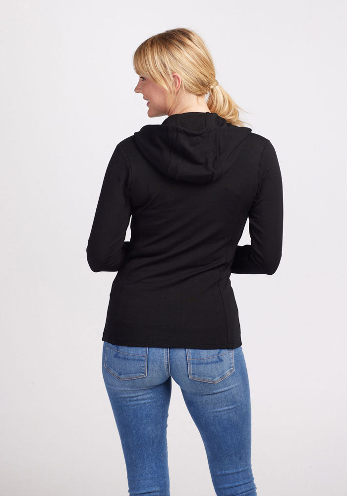 A person with blonde hair tied in a ponytail is standing with their back to the camera. They are wearing a Woolx Zoey Zip Up Hoodie in black and blue jeans. The background is plain and light-colored.